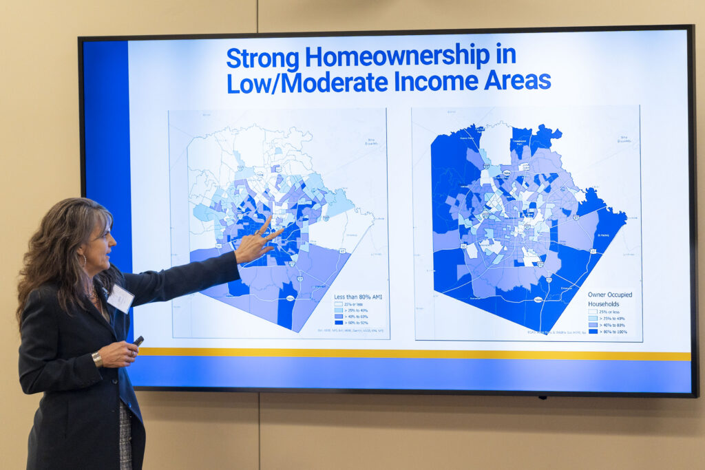 Laura McKieran, executive director of Community Information Now (CINow), presents research findings on heirs’ property in Bexar County. She stands in front of a large screen displaying maps and the heading “Strong Homeownership in Low/Moderate Income Areas,” gesturing toward the data while speaking. An American flag stands beside the presentation screen.